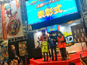 three boys standing on stage with judges at g1 beyblade tournament