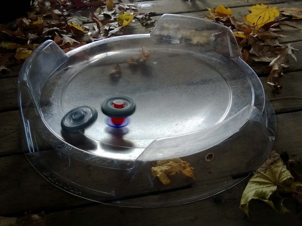 two beyblades battling in clear metal fight beyblade attack stadium surrounded by yellow leaves