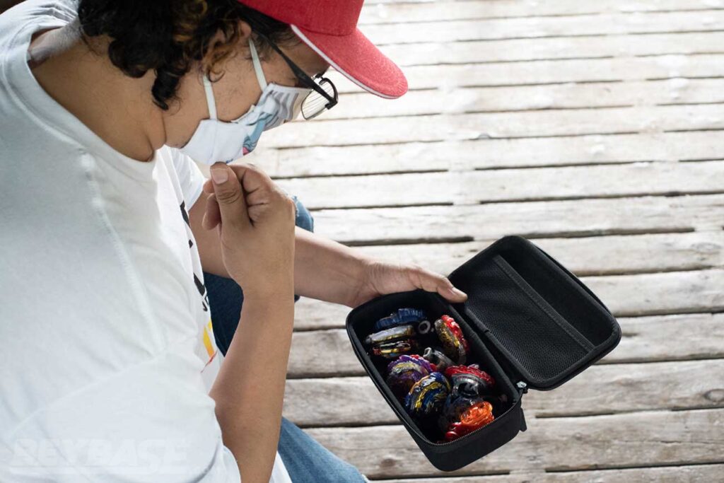 man wearing white shirt and red hat looking down into open black case filled with Beyblades