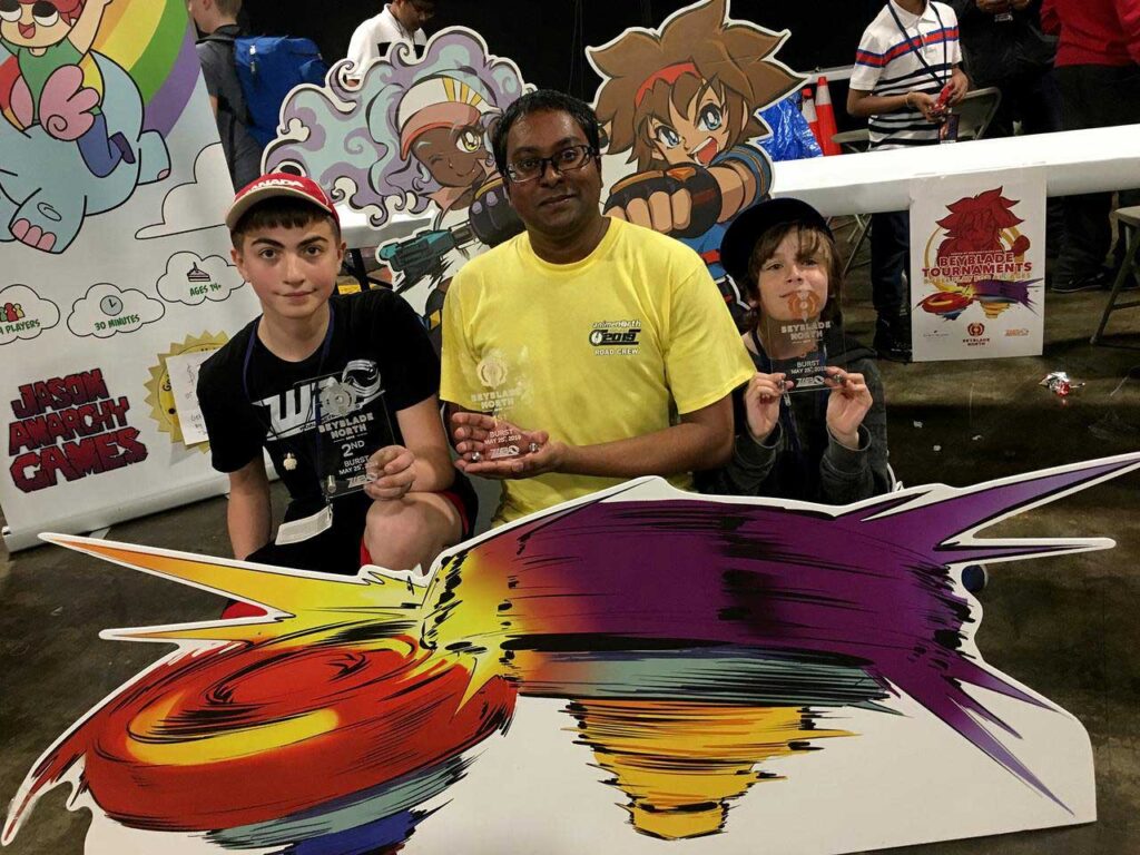 three people holding trophies between cutouts of world beyblade organization mascots taka and fumi and cutout of beyblades in the foreground