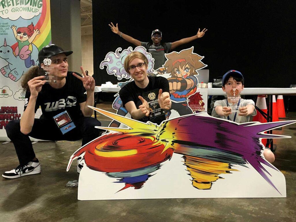 three people holding trophies between cutouts of world beyblade organization mascots taka and fumi and cutout of beyblades in the foreground