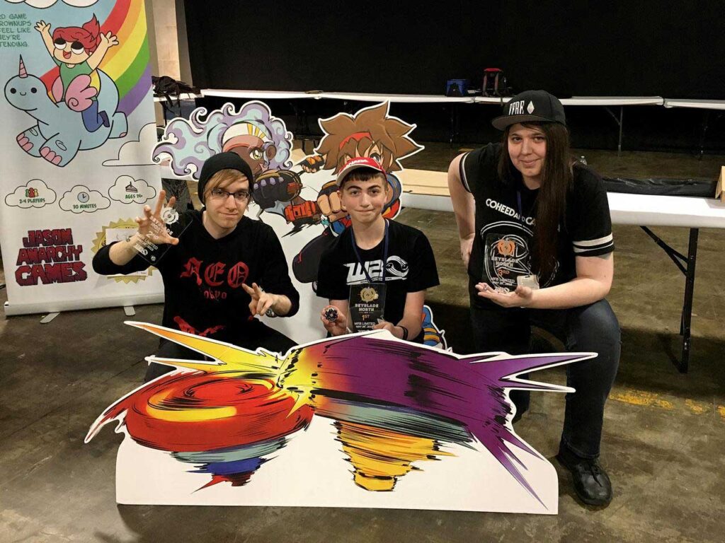 three people holding trophies between cutouts of world beyblade organization mascots taka and fumi and cutout of beyblades in the foreground